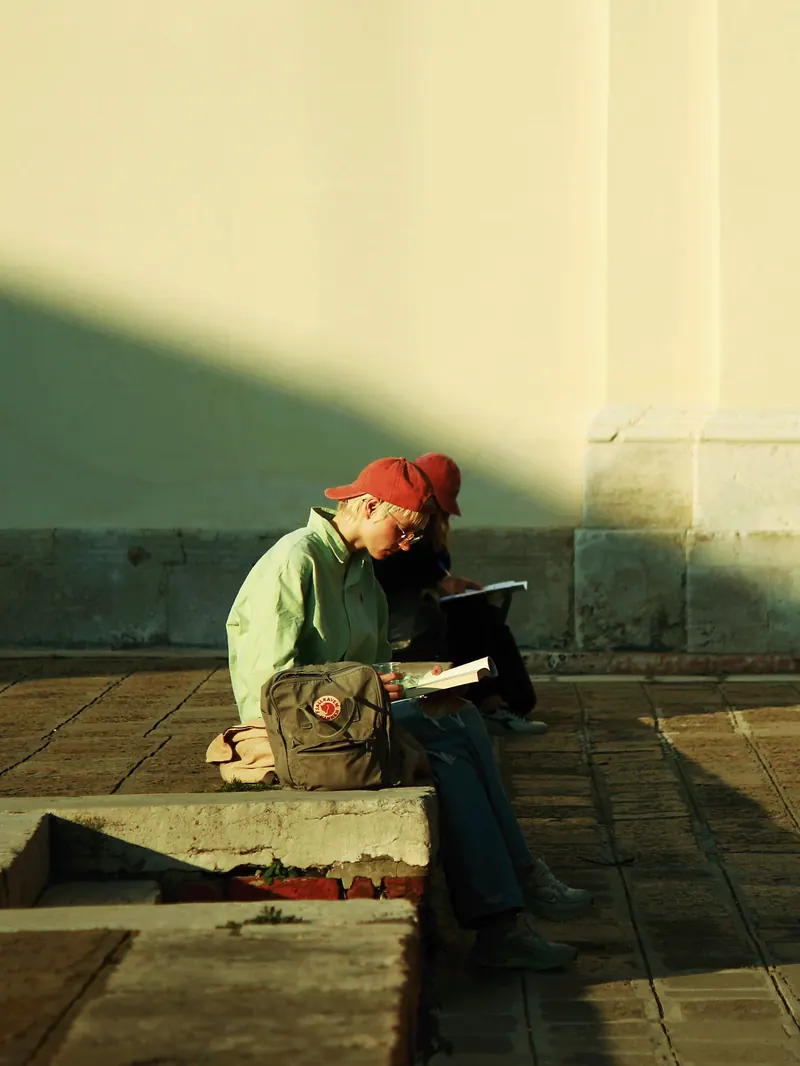 Hombre leyendo sentado en un escalón bajo una franja de sol.